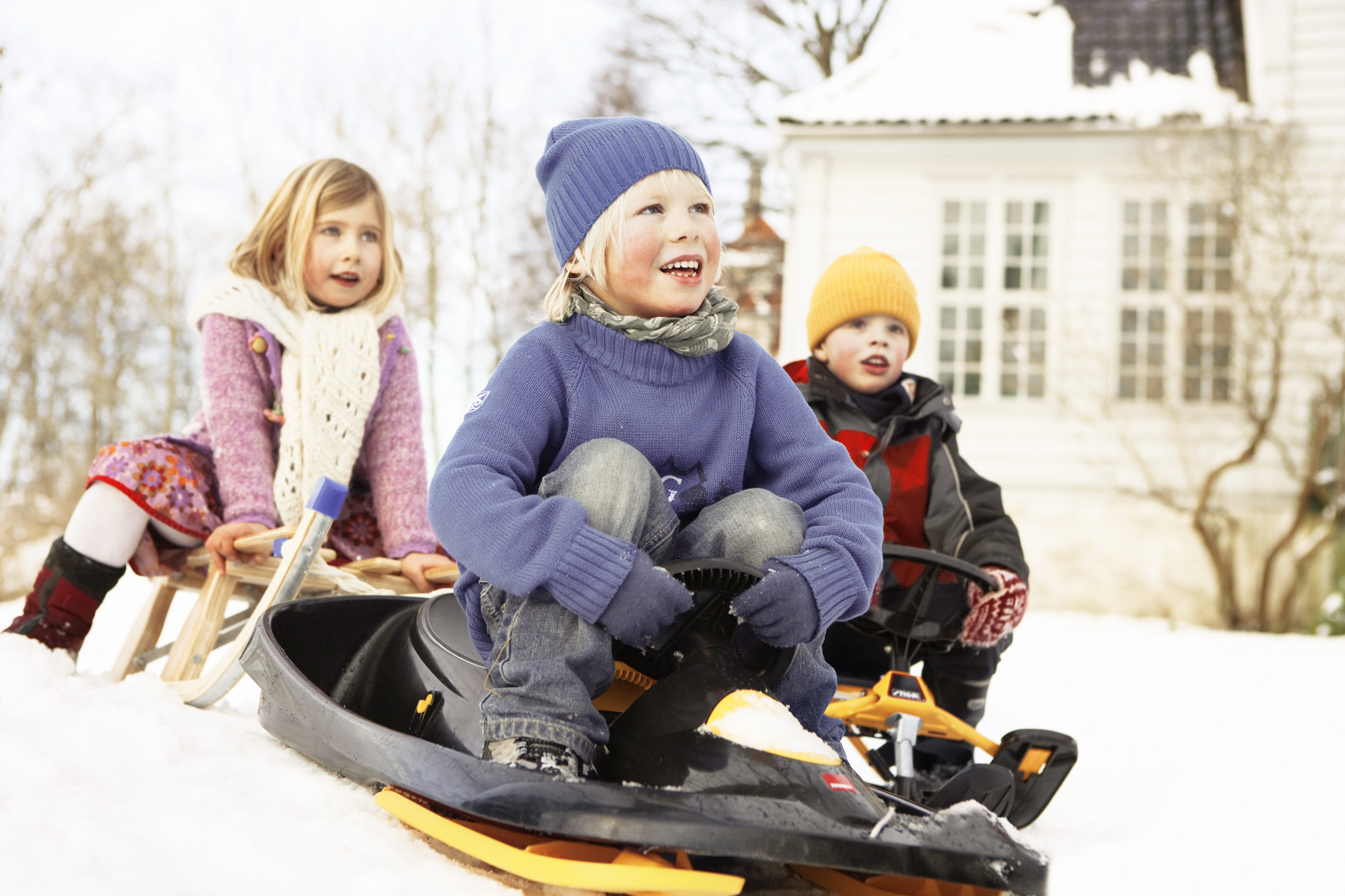 Tre barn sitter på kälkar, snö på marken