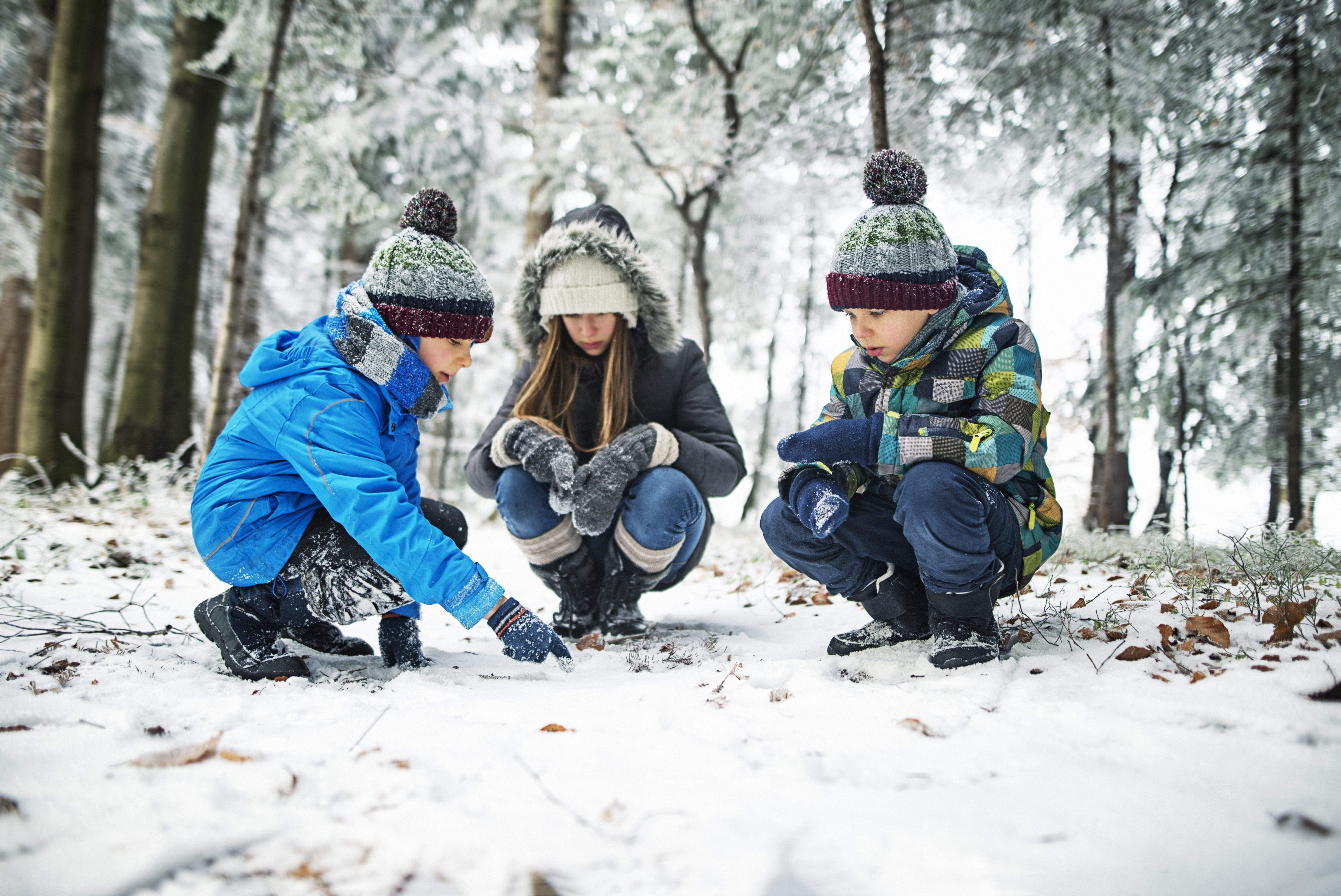 Tre varmt klädda barn sitter hukade vid en snötäckt mark