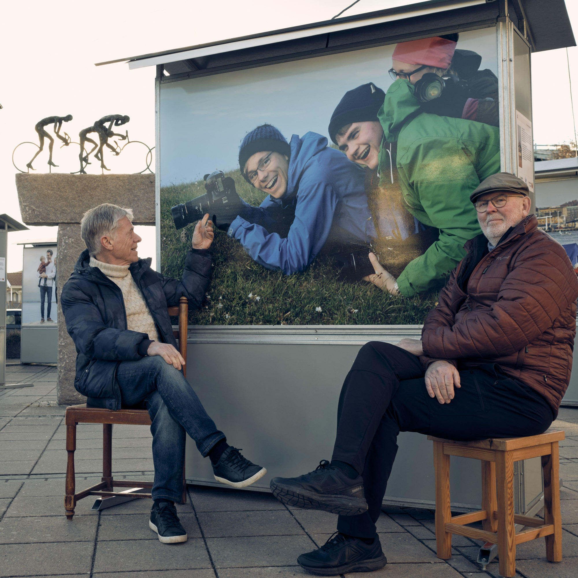 Två män sitter brevid ett stort foto på utomhusutställning av stor